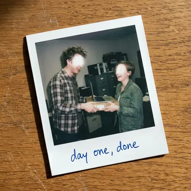 A Polaroid of two people in an office handing over a laptop, captioned in ballpoint: day one, done