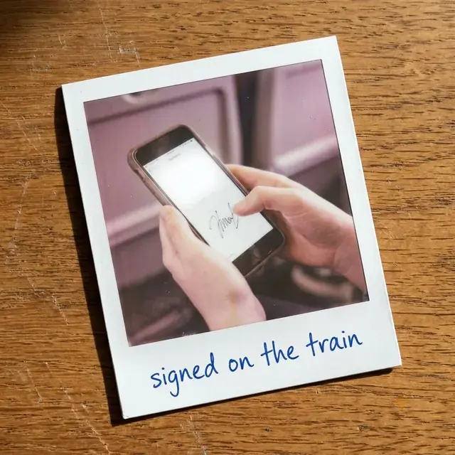 A Polaroid of someone signing on a phone screen, captioned in ballpoint: signed on the train