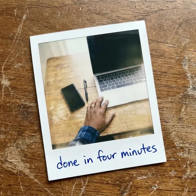 A Polaroid looking down at a desk with a laptop, a phone and a pen, captioned in ballpoint: done in four minutes